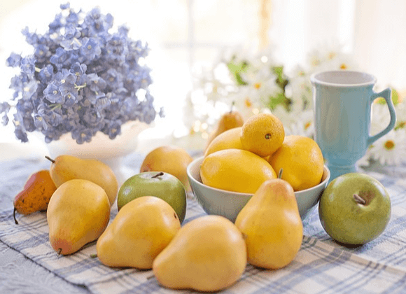 길쭉한 서양 배들이 여러개 있고, 뒤에 보라색꽃이 담긴 하얀 꽃병과 하늘색 머그컵이 있다.