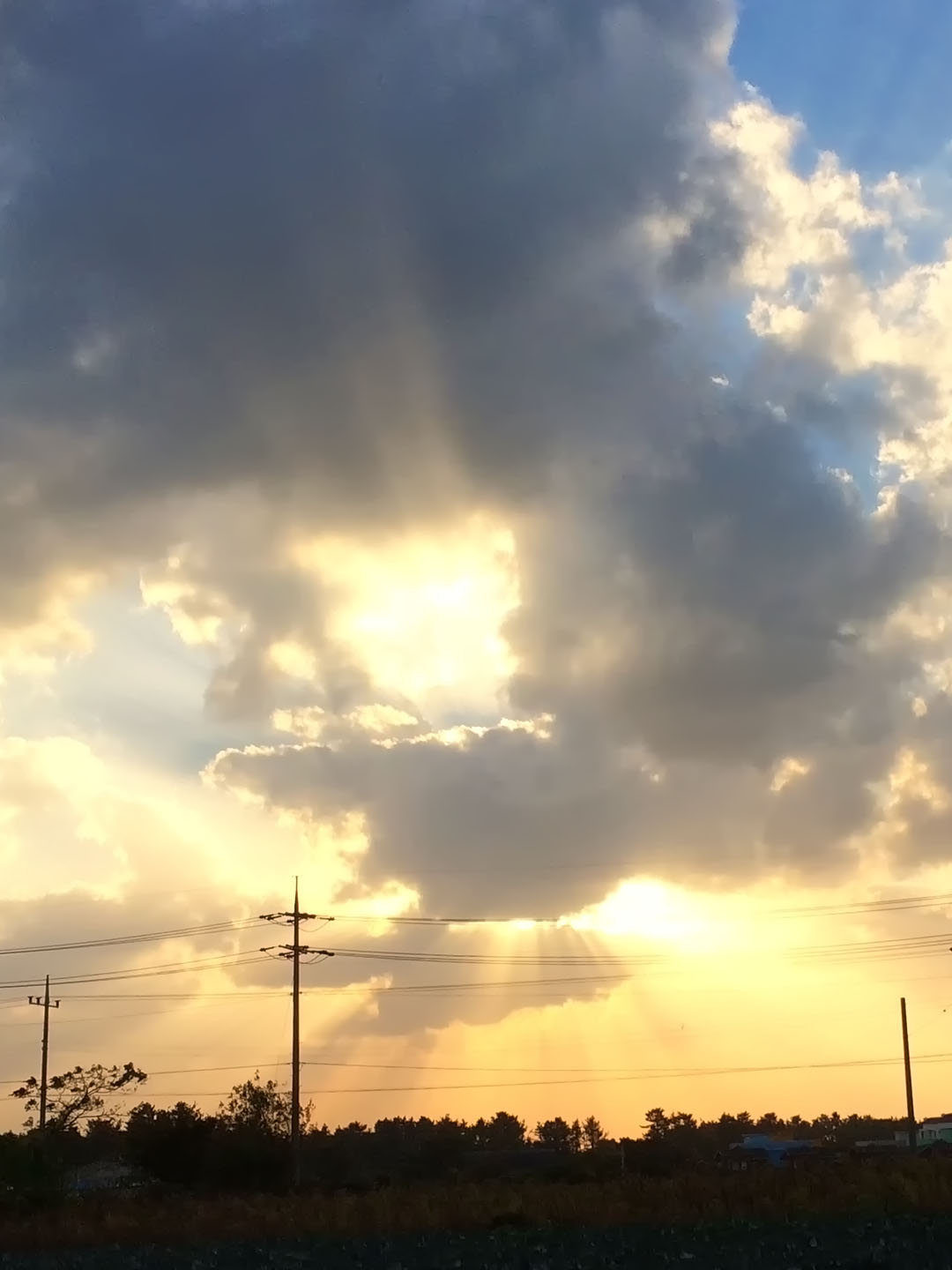 구름이 가득한 하늘 사이로 황금빛 햇살이 여러 갈래로 내려오며 들판과 전봇대 위로 빛내림이 펼쳐지는 평화로운 저녁 하늘 풍경 무료이미지