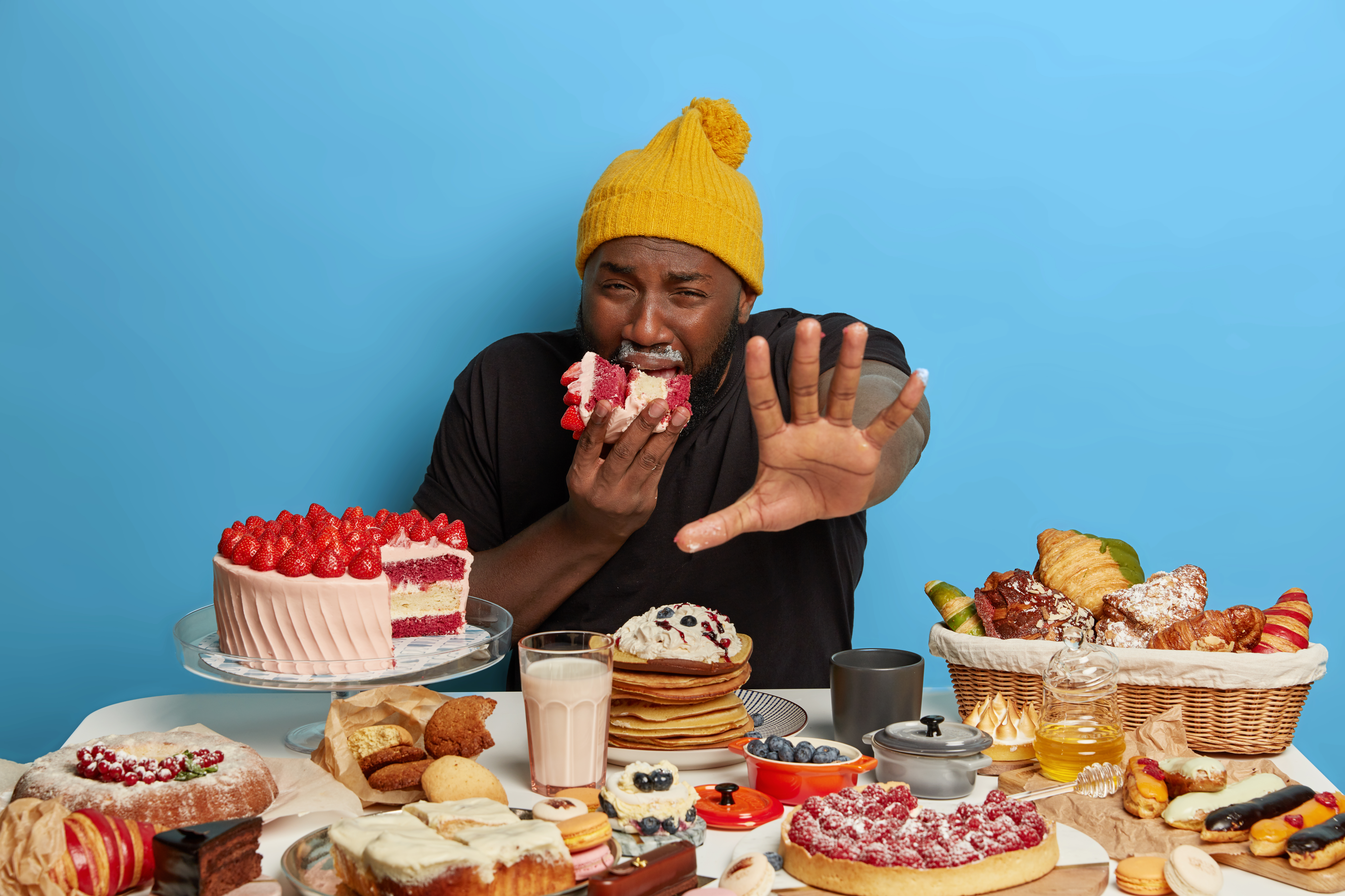 hungry-displeased-plump-afro-man-keeps-palm-forward-camera-bites-huge-piece-creamy-cake-gets-much-calories-surrounded-with-tasty-baked-products