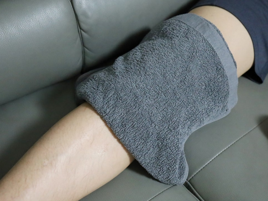 무릎이 아픈 사람이 쇼파에 앉아서 뜨거운 물에 담근 수건을 아픈 무릎 위에 놓아두고 찜질을 하고 있는 것을 찍은 사진
