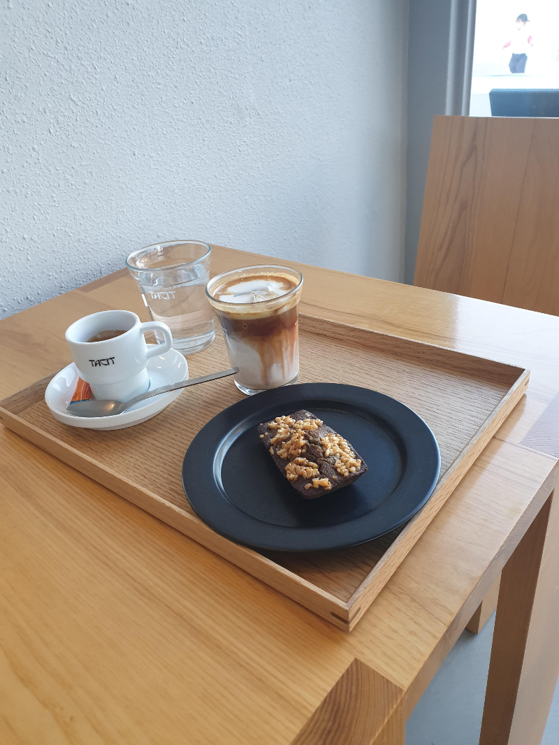 테이블 위에 트레이가 놓여 있고. 트레이 위에는 에스프레소, 라테, 물 그리고 휘낭시에가 놓여있다.