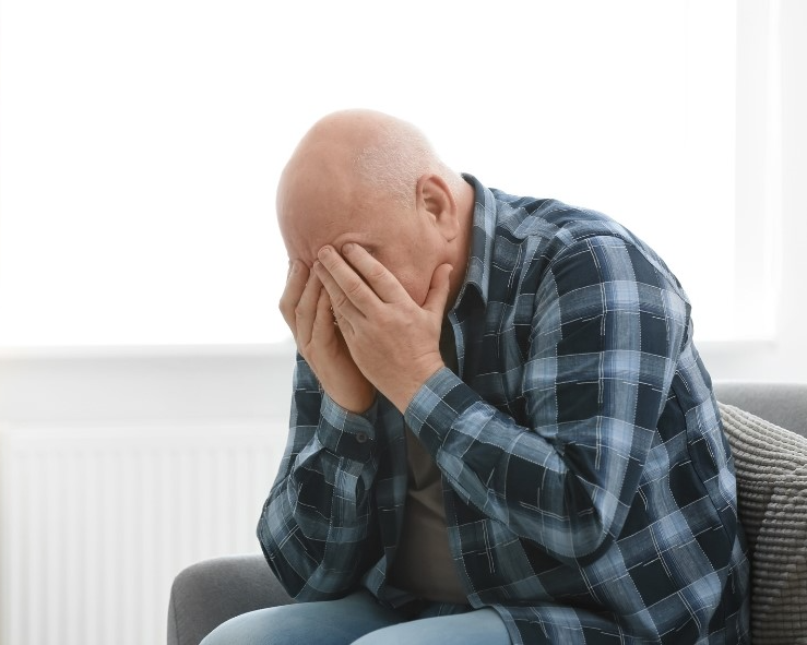 파란색 체크 셔츠를 입은 중년 남성이 소파에 앉아 두 손으로 얼굴을 감싸고 우울감에 빠져있는 모습 via canva