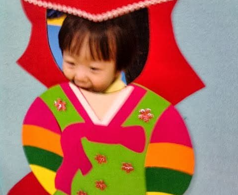 한복 모양으로 꾸민 포토존에 얼굴을 내밀고 웃고 있는 어린아이의 모습입니다.
알록달록한 색감의 전통 의상 장식과 아이의 자연스러운 표정이 어우러져 설날 특유의 따뜻하고 정겨운 분위기를 전합니다.