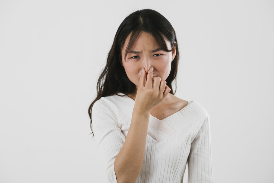 하얀색 니트를 입은 젊은 동양 여성이 방귀 냄새 때문에 손으로 본인의 코를 잡고 냄새를 억제하고 있는 것을 찍은 사진