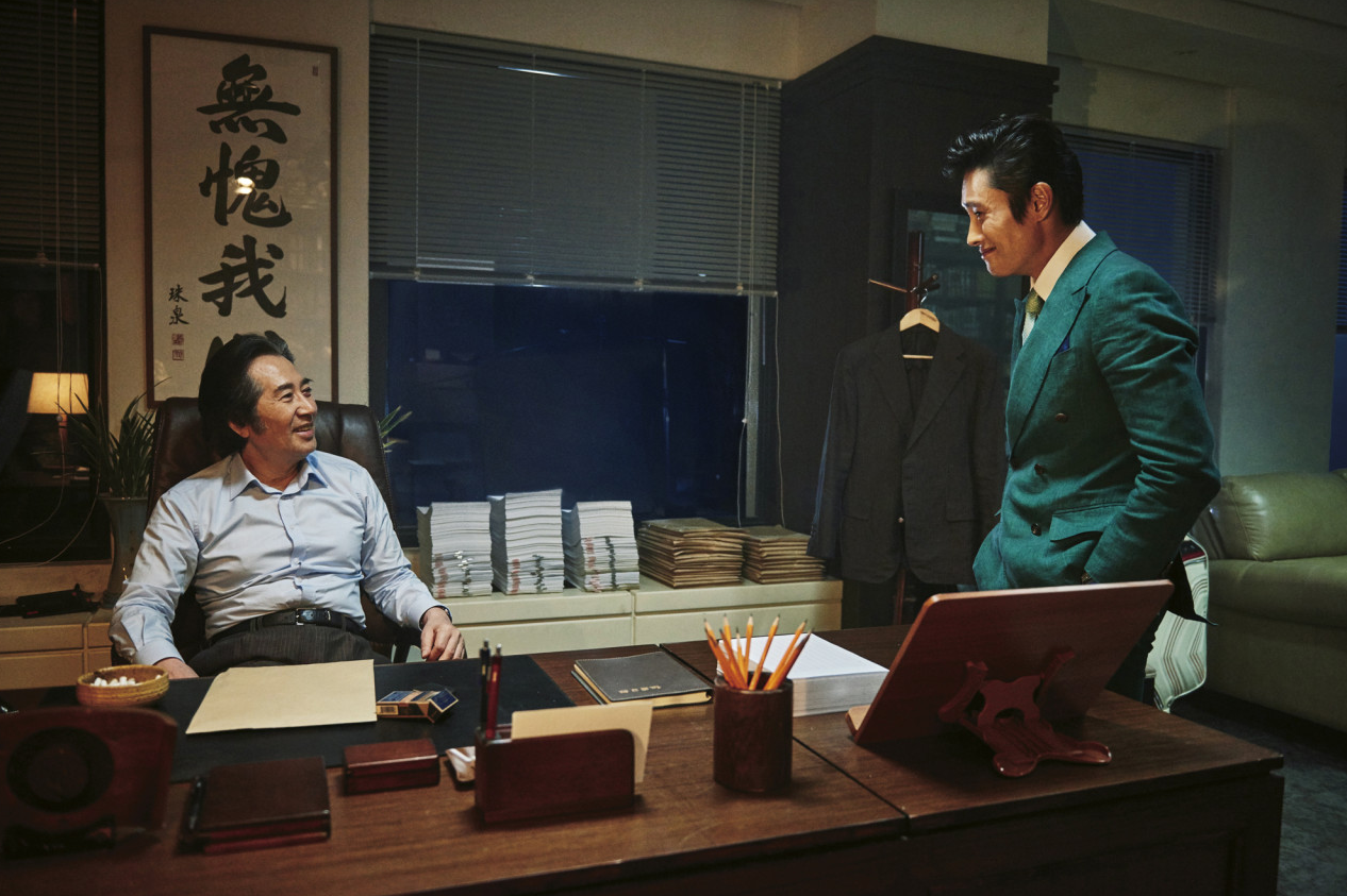 어두운 사무실 안에서 책상에 현금 다발을 쌓아두고 이강희와 마주한 장면, 뒤로 한자 액자가 걸려 있다