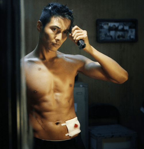 영화 &lsquo;아저씨&rsquo;의 한 장면. 배우 원빈이 상반신을 드러낸 채 거울 앞에서 전기 이발기로 스스로 머리를 밀고 있다. 복부에는 피가 스며든 붕대가 붙어 있으며, 배경은 어두운 실내 공간이다.