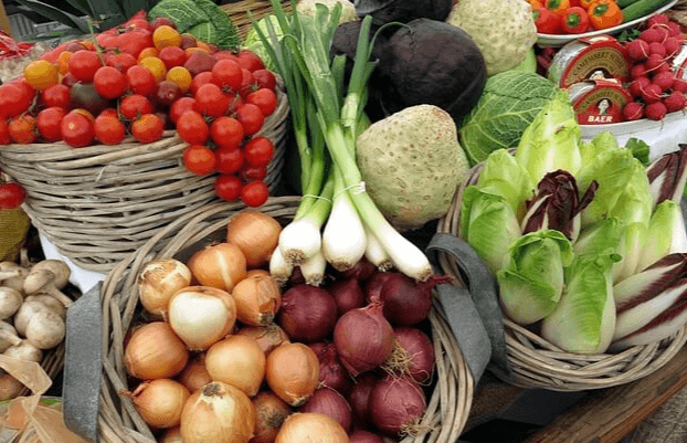 갈색 바구니에 양파가 한가득 있고, 작은 배추들도 옆 바구니에 가득하다. 뒤에는 바구니에 빨간 토마토가 가득하고, 맨 위에는 대파 한묶음이 놓여있다.