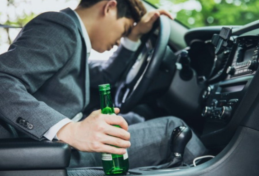 한 동양의 젊은 남성이 술을 마신 후에 자동차 운전석에 앉아서 한쪽 손에는 소주병을 쥐고, 운전대에 머리를 박고 자고 있는 것을 찍은 사진