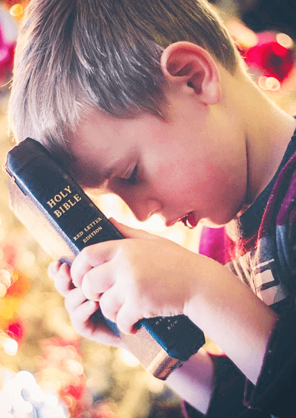 짧은 갈색 머리를 가진 백인 남자 아이가 두 눈을 감고 두 손으로 검정색 성경을 들고 이마에 대고 있다.