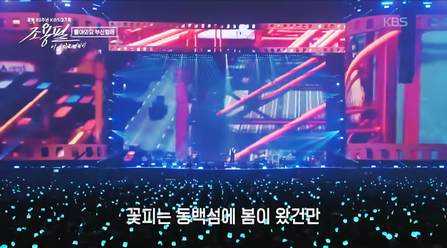 ‘꽃피는 동백섬에 봄이 왔건만’ 가사 자막이 떠 있는 무대 전경. 푸른 조명 속 관객들의 응원봉이 바다처럼 빛나는 장면.