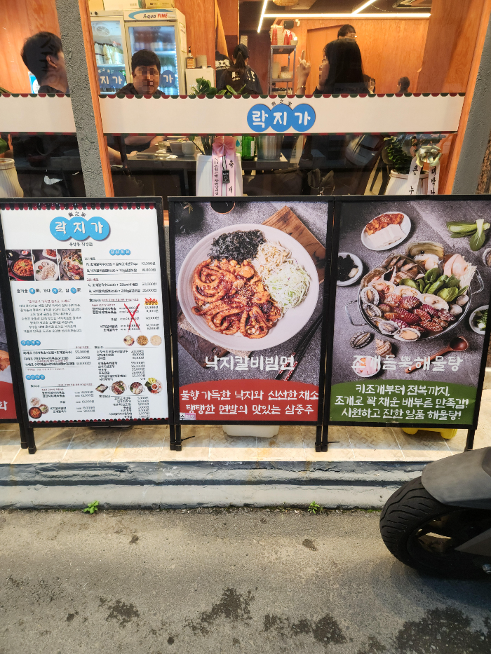 락지가의 건물 바깥에 락지가의 메뉴와 대표메뉴인 낙지칼비빔면과 조개듬뿍해물탕이 그려진 입간판이 있다