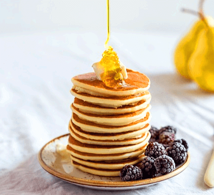 접시에 12장의 동그란 팬케이크가 쌓여있고 맨 위에는 금색 꿀이 떨어지고있다.