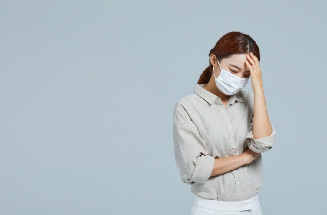 한 여자가 마스크를 쓰고 손으로 머리를 만지며 괴로워하고 있다.