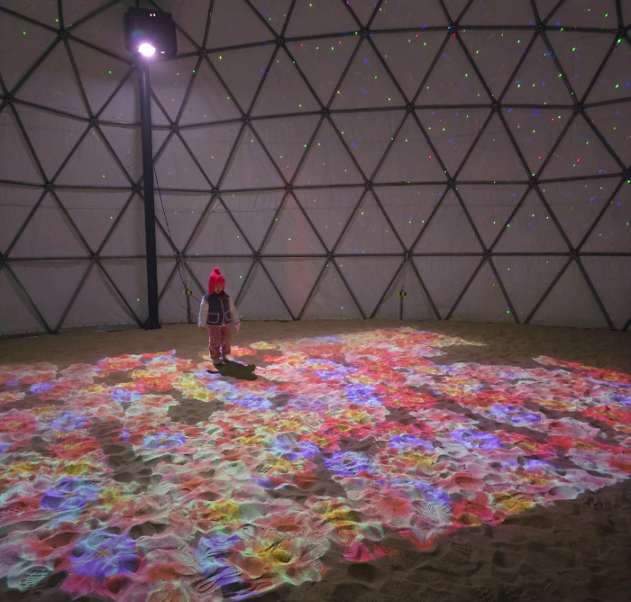 빛으로 쏘아 만든 모래 위 꽃그림 위를 아기가 걷고있다.