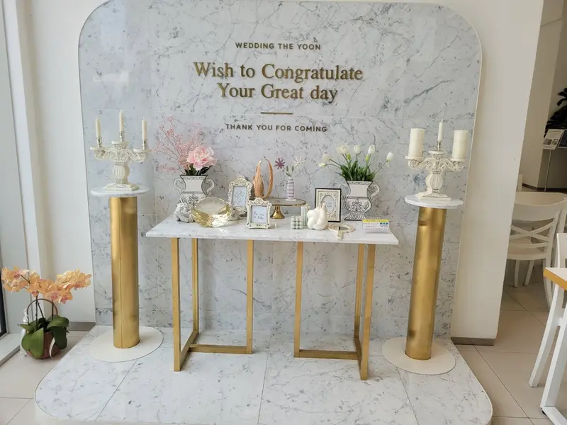 해그리다카페 웨딩홀의 대리석 포토존과 골드 장식, 꽃과 촛대, 환영 메시지가 어우러진 세련된 웨딩 데코 공간