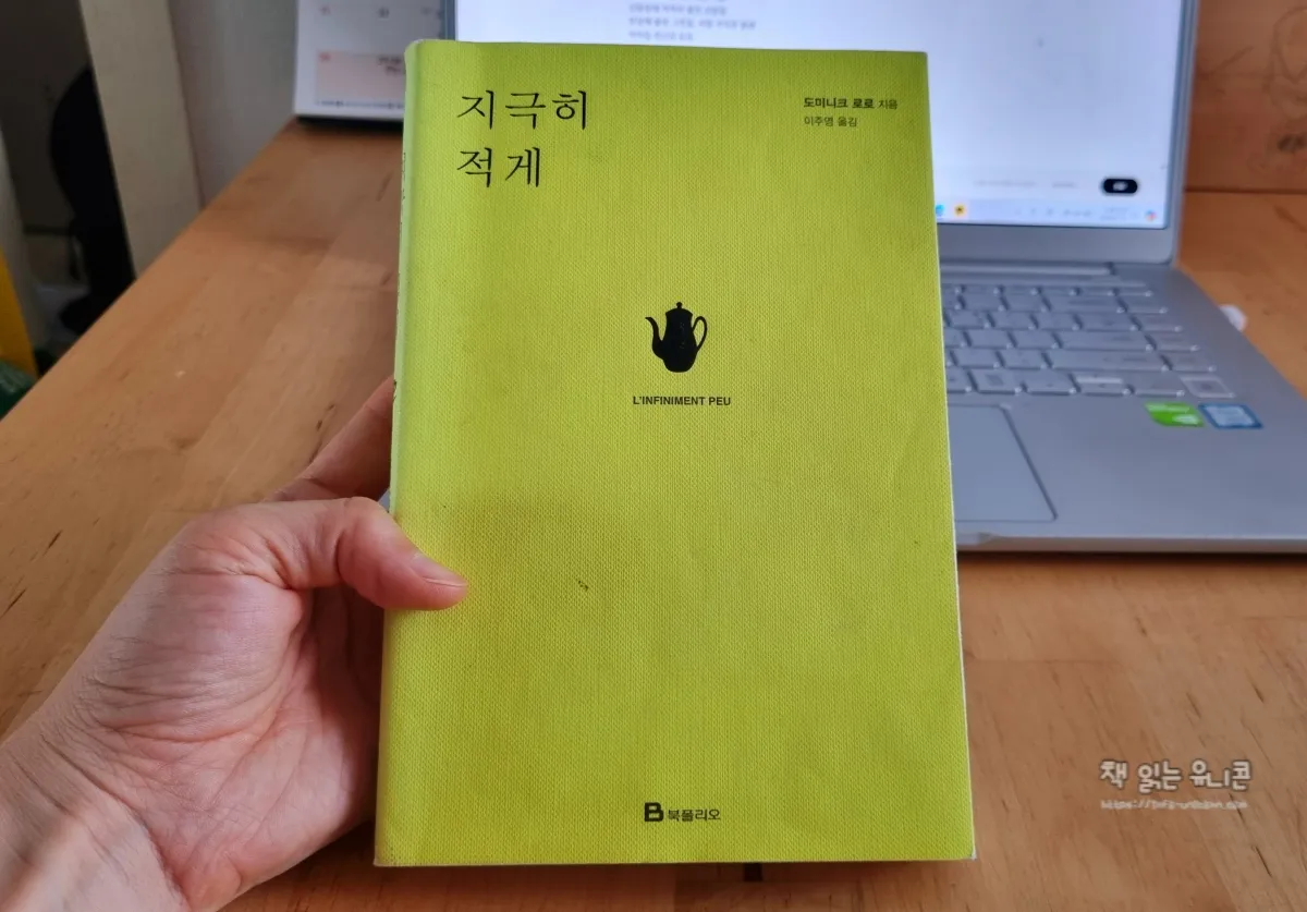 지극히 적게 라는 제목의 책을 표지가 잘보이게 손으로 들고 있다. 뒤에 노트북이 놓여있다.