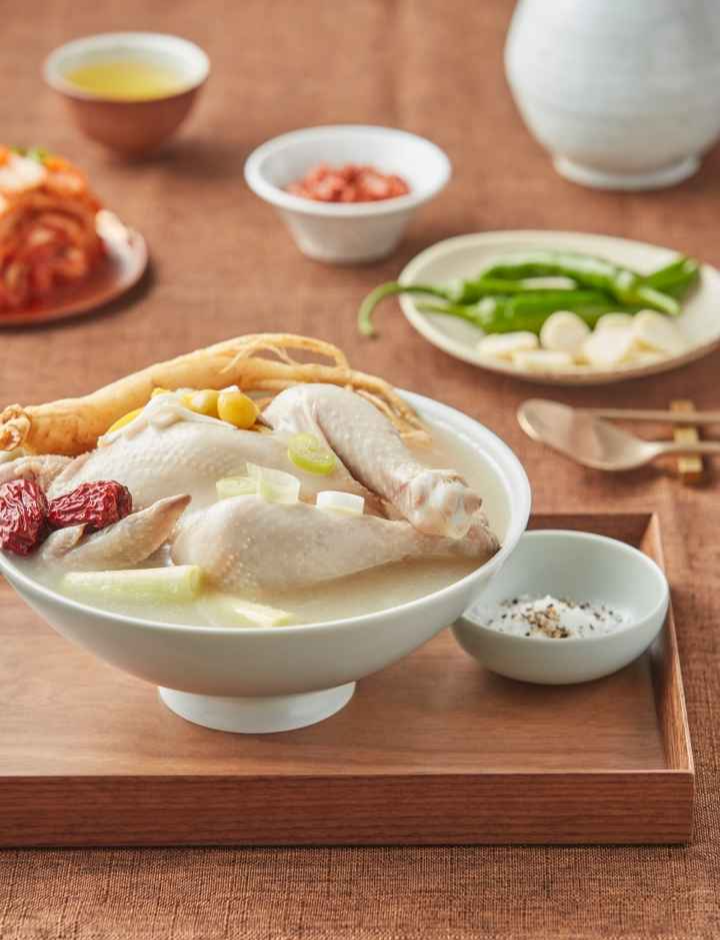 A traditional Korean dish of Samgyetang (ginseng chicken soup) served in a white bowl with ginseng root, jujube, garlic, and green onion. Side dishes like kimchi, green chili peppers, and salt with pepper are visible in the background.