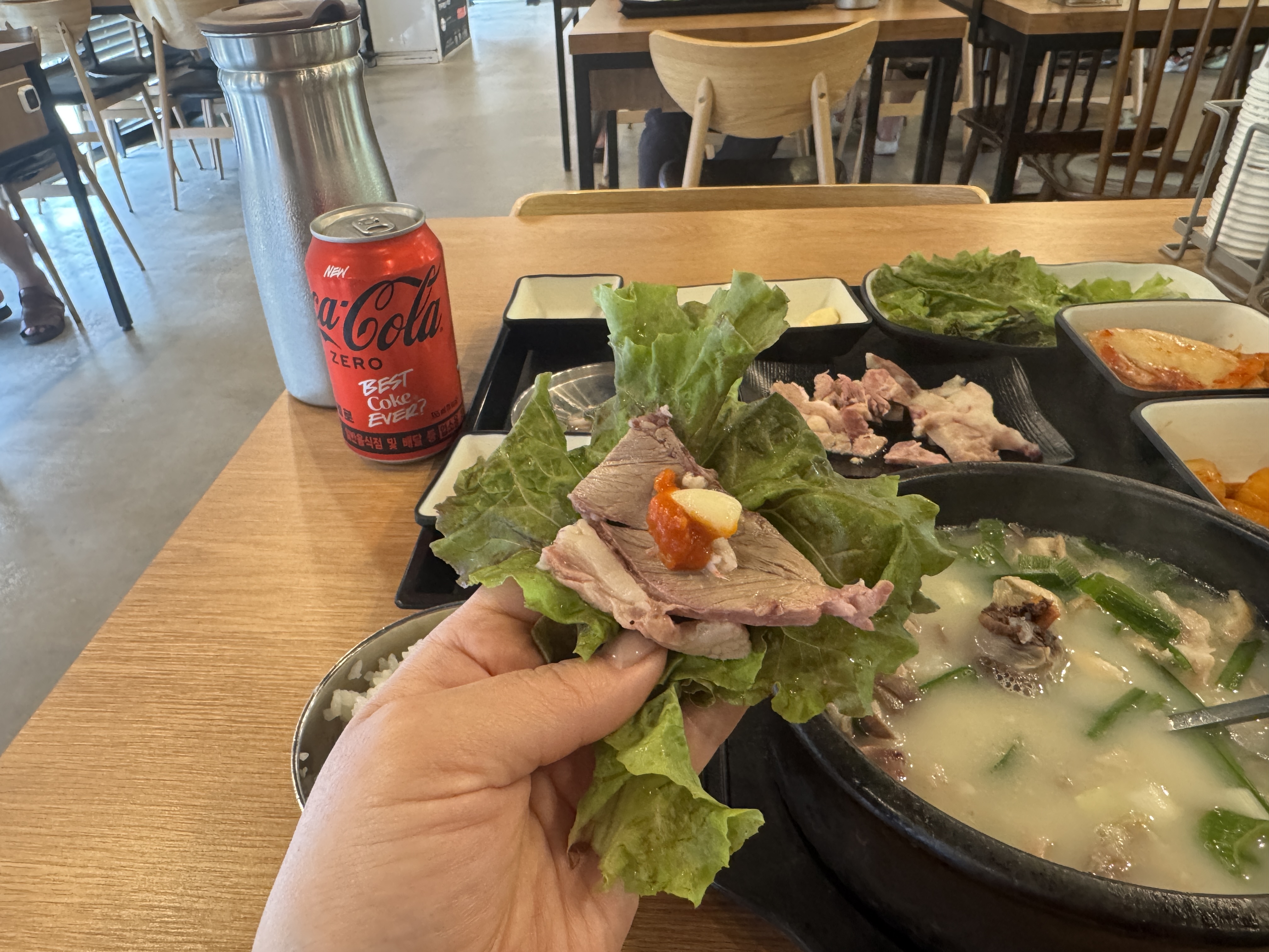 경자국밥 용인본점에서 쌈을 싸는 장면. 상추 위에 수육과 마늘, 쌈장이 올려져 있으며, 테이블에는 끓고 있는 국밥 뚝배기와 밑반찬, 콜라 제로 캔이 놓여 있다.