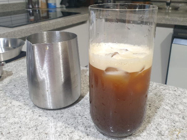 테이블 위에 스팀피처와 유리잔이 놓여있다. 유리잔 안은 거품과 커피가 나눠져 있다.