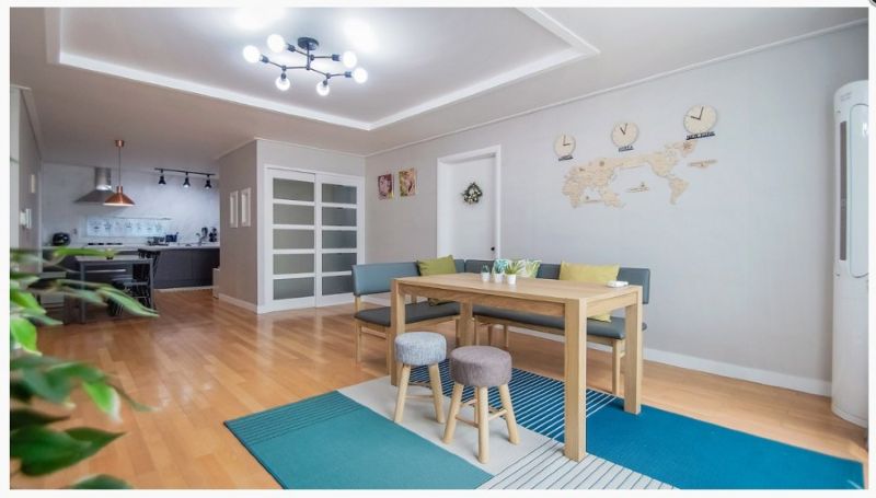 넓고 깨끗한 우드 톤 바닥의 쉐어하우스 거실. 디자인 식탁 세트와 세련된 블랙 펜던트 조명, 벽면의 세계지도 인테리어가 조화로운 모습