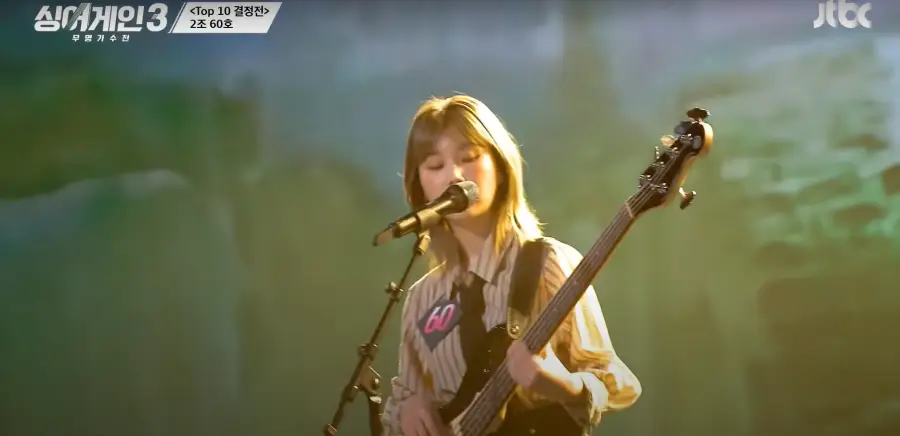 싱어게인 3- 초록색을 배경으로 가슴에 60번호표를 달고 세로줄무늬 상의를 입고 단발머리를 늘어뜨린채 기타를 보며 노래하는 60호 상반신