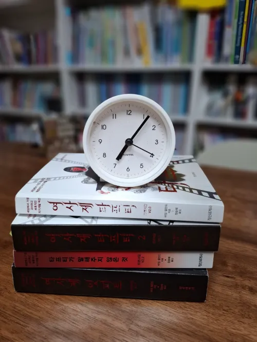 여사제 타프티 책 위에 놓인 동그란 탁상시계 사진 - 거울의 지연 현상과 시간의 흐름을 상징함 (A photo of a round desk clock on the Tufti the Priestess book, symbolizing the mirror delay and the flow of time)