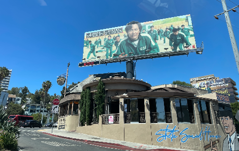 건물 위로 큰 광고판이 있다. 미국에서 크게 흥행한 우리나라의 넷플릭스 드라마, 오징어 게임의 광고이다. 주인공인 이정재 배우가 가운데 있고 그 뒤로 다른 배우들이 있다. 모두 초록색 트레이닝복을 입고 있다.