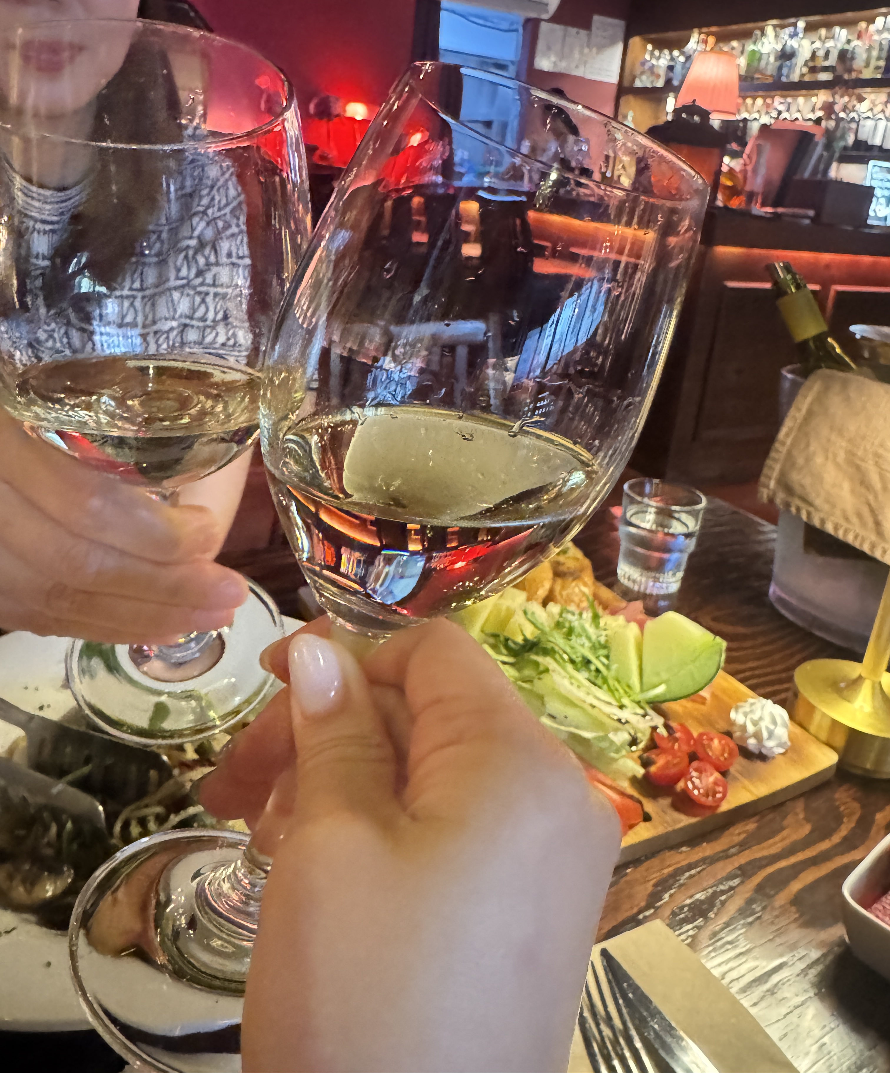 논현동 비노비스 와인바에서 두 사람이 화이트 와인잔을 들고 조심스럽게 짠을 하는 장면. 테이블 위에는 하몽 플레이트와 안주들이 놓여 있고, 배경에는 아늑한 조명의 바가 보인다.