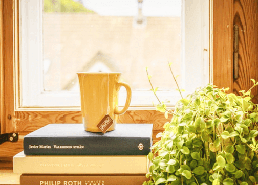 창가 선반에 책 3권이 쌓여있고 책 위에는 차가 담긴 노란 컵이 있다. 그리고 오른쪽에는 푸른 식물이 있다.