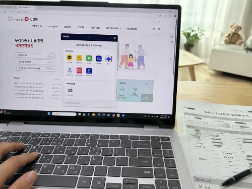 노트북 화면에 띄워진 국민건강보험공단 건강iN 홈페이지의 간편인증 로그인 팝업창과 키보드 위의 손, 노트북 오른편에 놓인 영유아 건강검진 결과통보서 종이 서류와 볼펜 사진