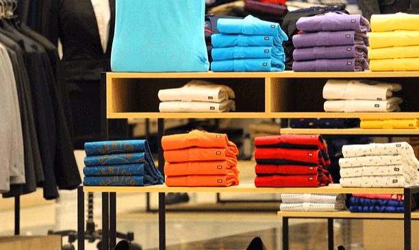 백화점 매장에 남자 셔츠와 옷들을 판매하는 진열대가 있다. 파란색, 주황색, 빨간색, 흰색 셔츠가 있다.