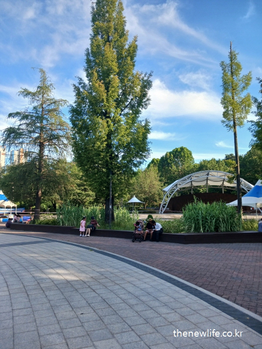 Families resting near the outdoor stage in Children's Grand Park, shaded by tall trees./서울 어린이대공원 야외무대 주변 벤치와 그늘에서 쉬는 가족들