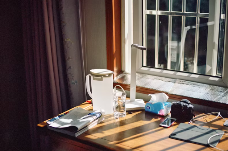 창가 햇살이 비치는 책상 위에 노트, 물컵, 전등이 놓여 있는 조용한 아침 작업 공간