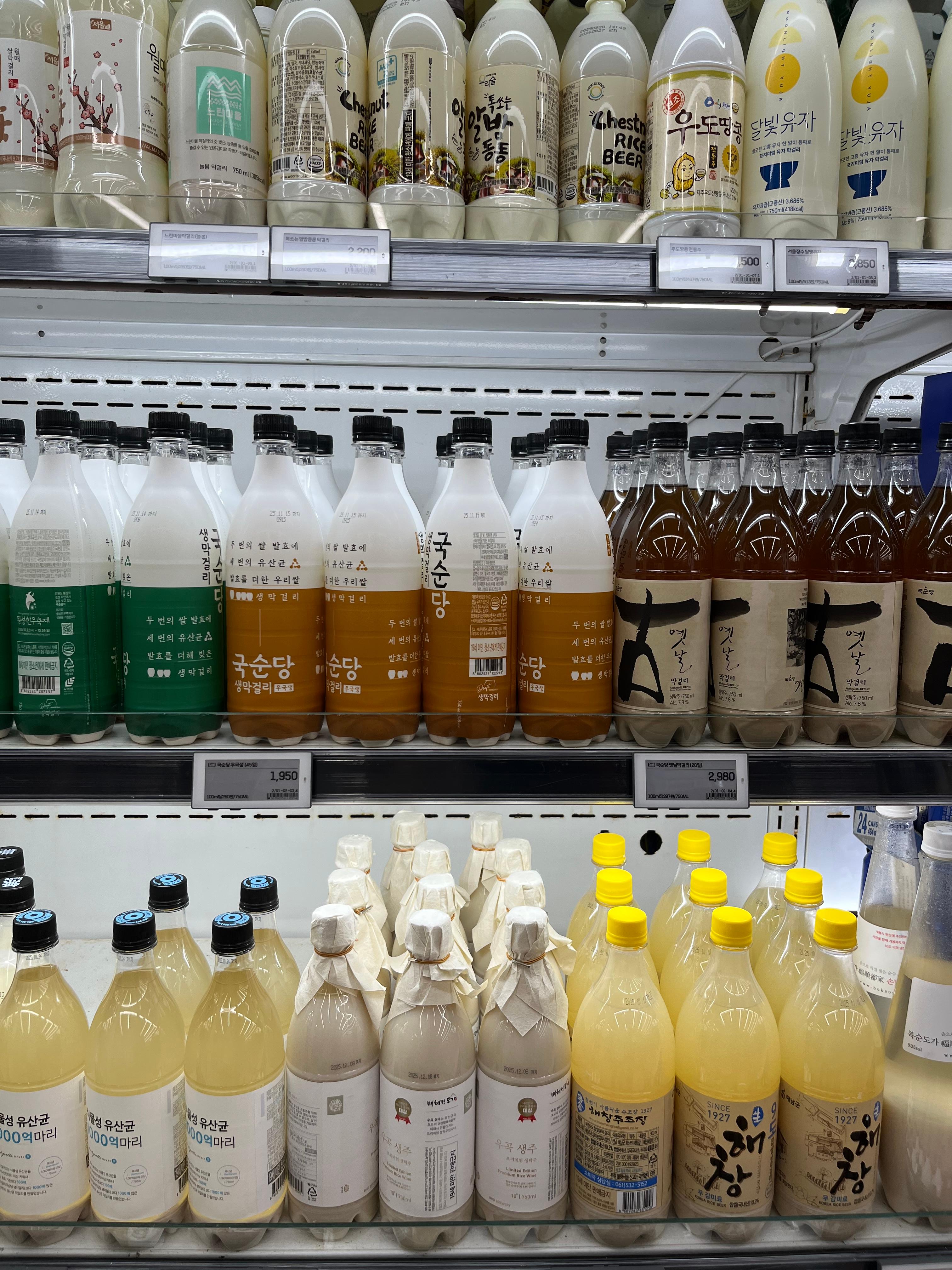 A refrigerator shelf in a Korean store displays various bottles of makgeolli &mdash; traditional Korean rice wine &mdash; in flavors like chestnut, citron, and plain rice, showing the modern diversity of Korea&rsquo;s oldest fermented drink.