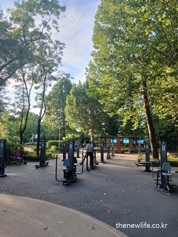 leg exercise machines in a shady park area with trees and people exercising/나무가 많은 그늘진 공원 속에서 하체 운동기구를 이용하는 시민들의 모습