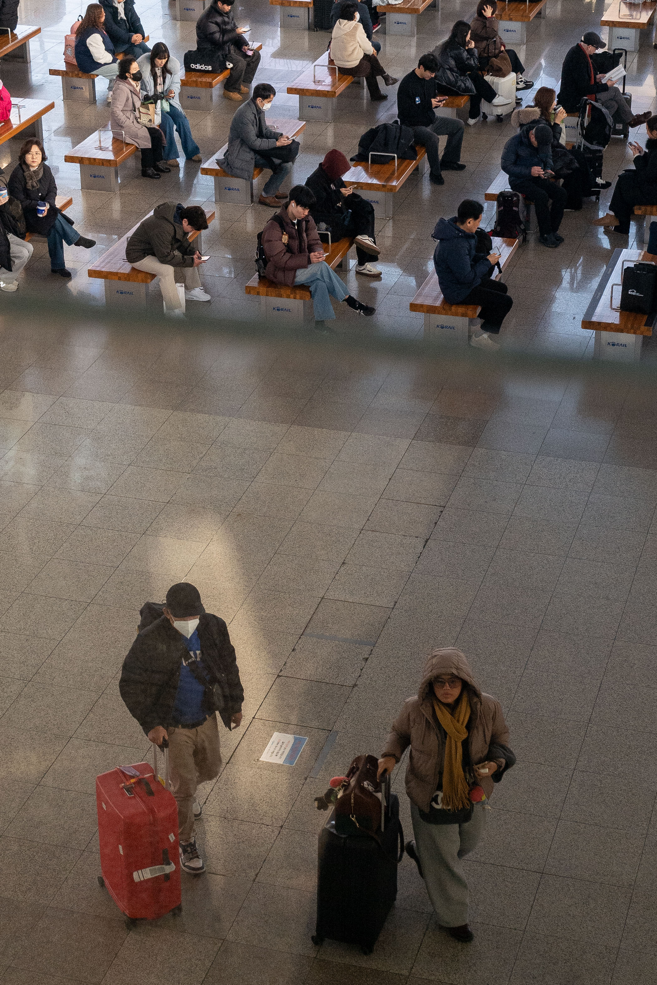 기차역 안. 사진의 1/3에는 균일하게 배열되어 있는 의자 위에 사람들이 빽빽하게 앉아있다. 그 아래 나머지 2/3에는 단 두 명의 사람이 캐리어를 끌고 가고 있는 모습이 대비된다.