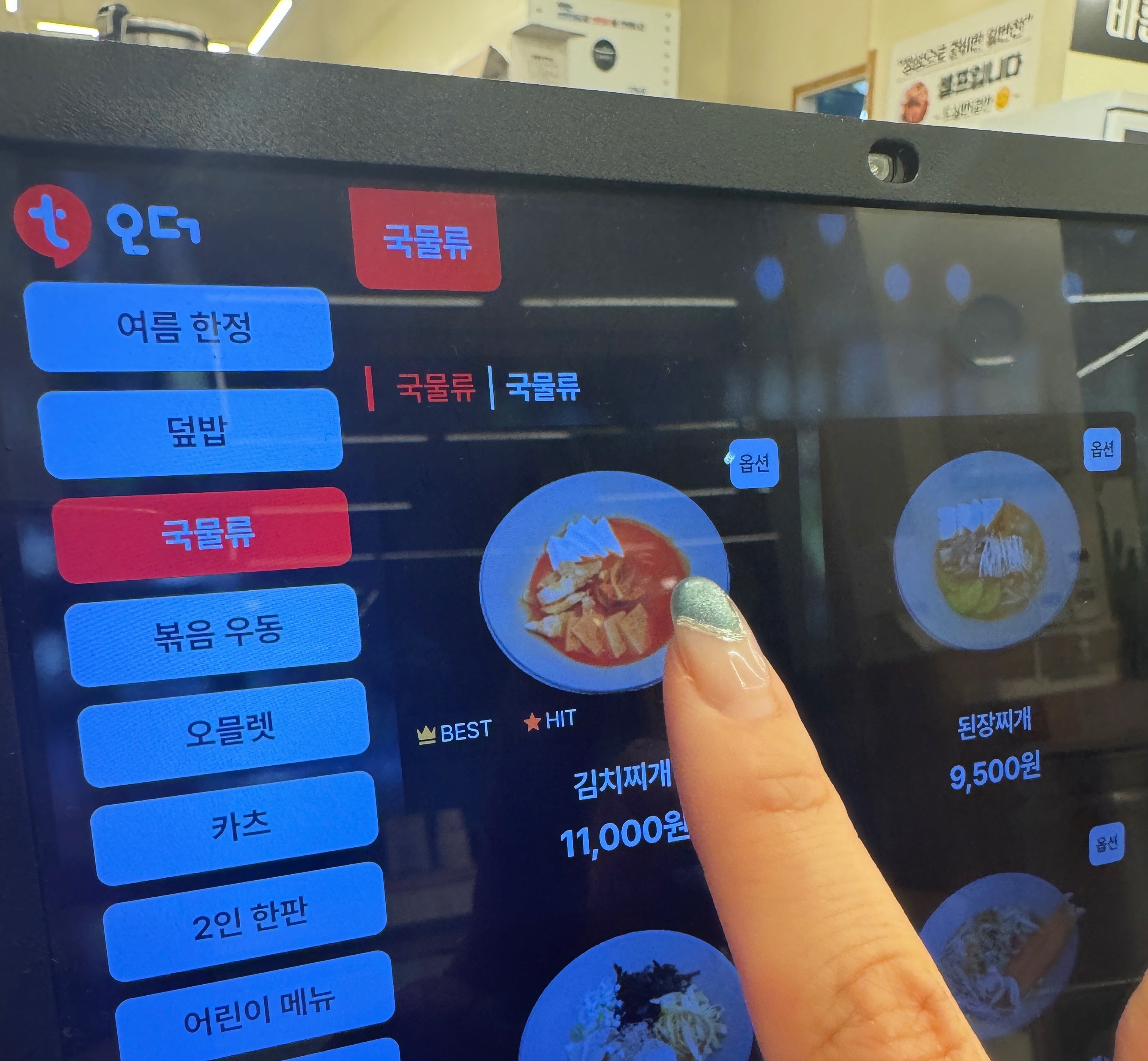용인 수지구 동천동 바른식탁 키오스크 화면. 손가락으로 김치찌개(11&amp;#44;000원) 메뉴를 선택하는 모습. 국물류 메뉴 중 김치찌개가 BEST&middot;HIT 표시로 강조되어 있다.