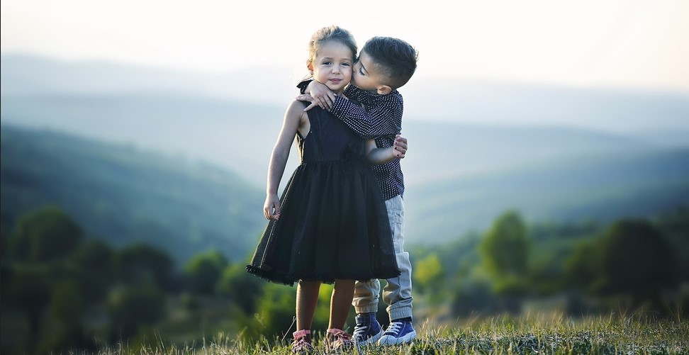 멀리 산이 보이는 아름다운 들판에서 어린 남자아이가 어린 여자아이 볼에 뽀뽀를 하는 사진