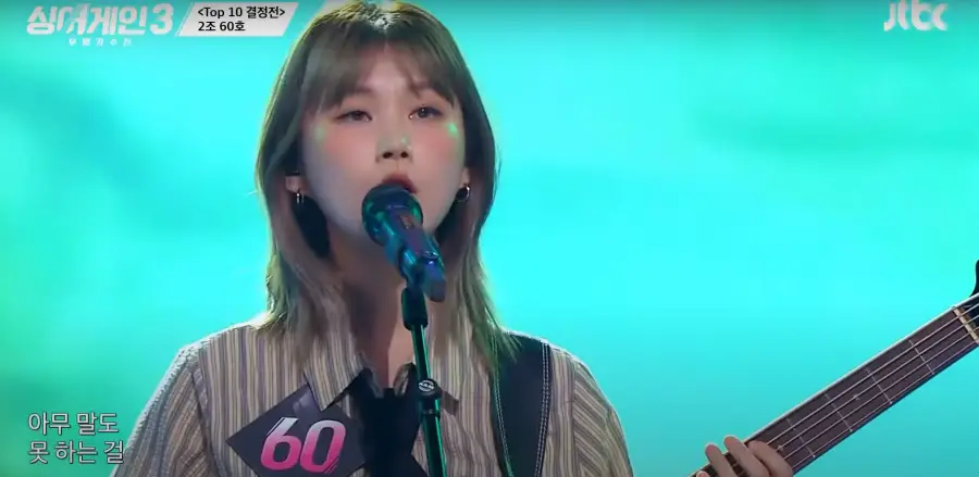 싱어게인 3- 청록색을 배경으로 가슴에 60번호표를 달고 세로줄무늬 상의를 입고 단발머리를 늘어뜨린채 기타를 치며 노래하는 60호 상반신