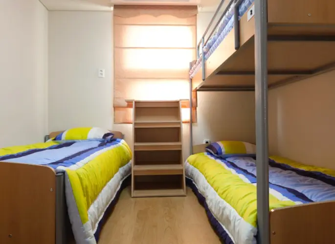 재수학원- 흰색 벽지 정면 커텐으로 가려진 밝은 창문 밑 갈색 수납장 좌측 1인침대 마루 통로 우측 2층 침대