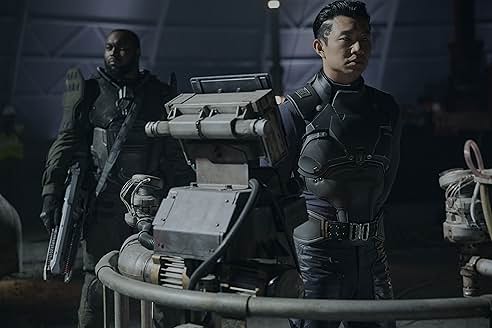 검은 전투복을 입은 두 인물이 미래형 장비 옆에 서 있는 장면