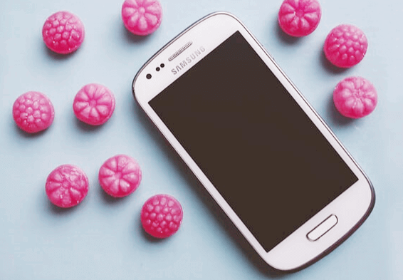 하얀색 삼성 갤럭시 핸드폰이 있고 주변에는 분홍색의 동그란 알사탕들이 깔려있다.