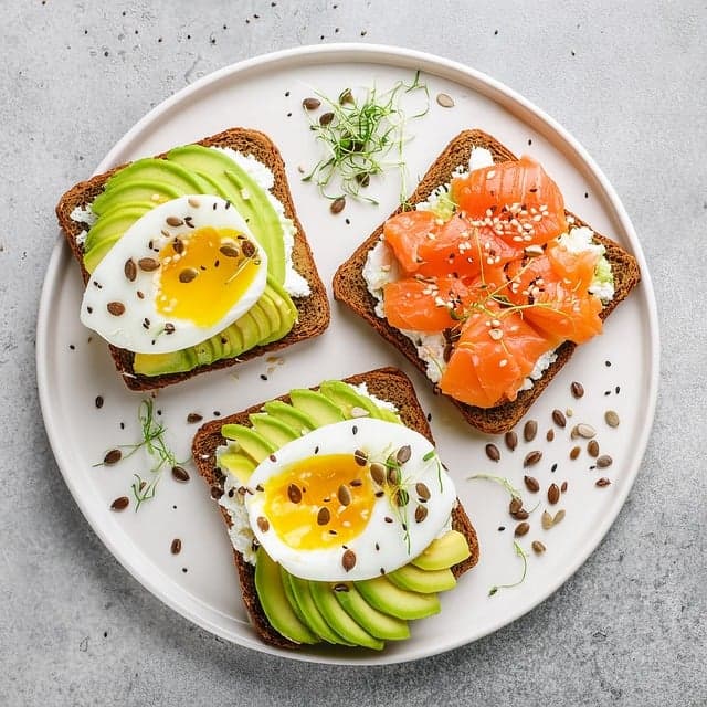 아보카도 슬라이스와 반숙 달걀을 올린 토스트 2조각, 훈제 연어 토스트 1조각이 흰 접시에 담긴 건강식