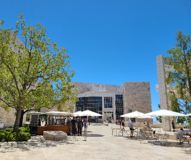 밝은 색의 건물이 동서남북으로 있고&amp;#44; 가운데에는 흰 파라솔과 테이블&amp;#44; 벤치가 있다. 