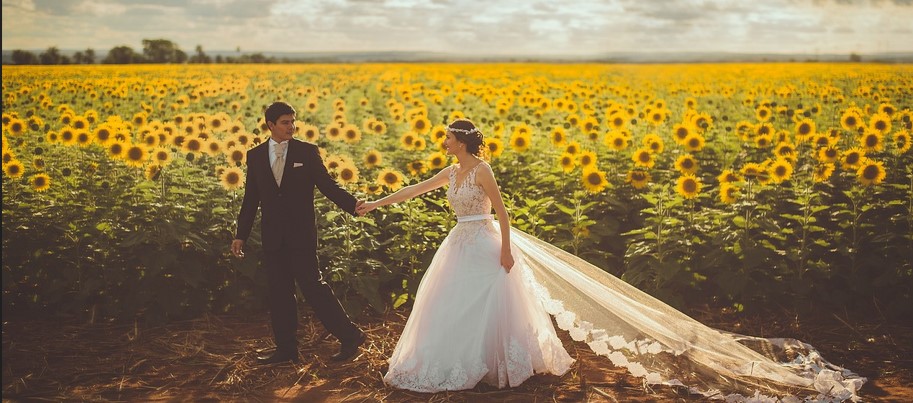 해바라기가 가득 피어있는 들판에 방금 결혼식을 올린듯한 신랑 신부가 양복과 드레스를 입고 손을 맞잡은채 걷고 있는 사진
