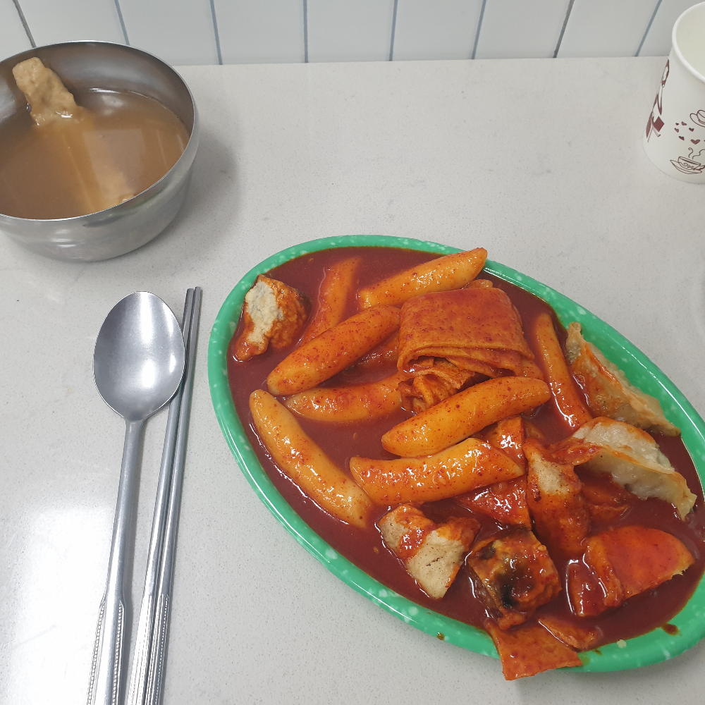 흰 테이블 위에 떡볶이와 어묵 국물 그리고 수저가 놓여있다.