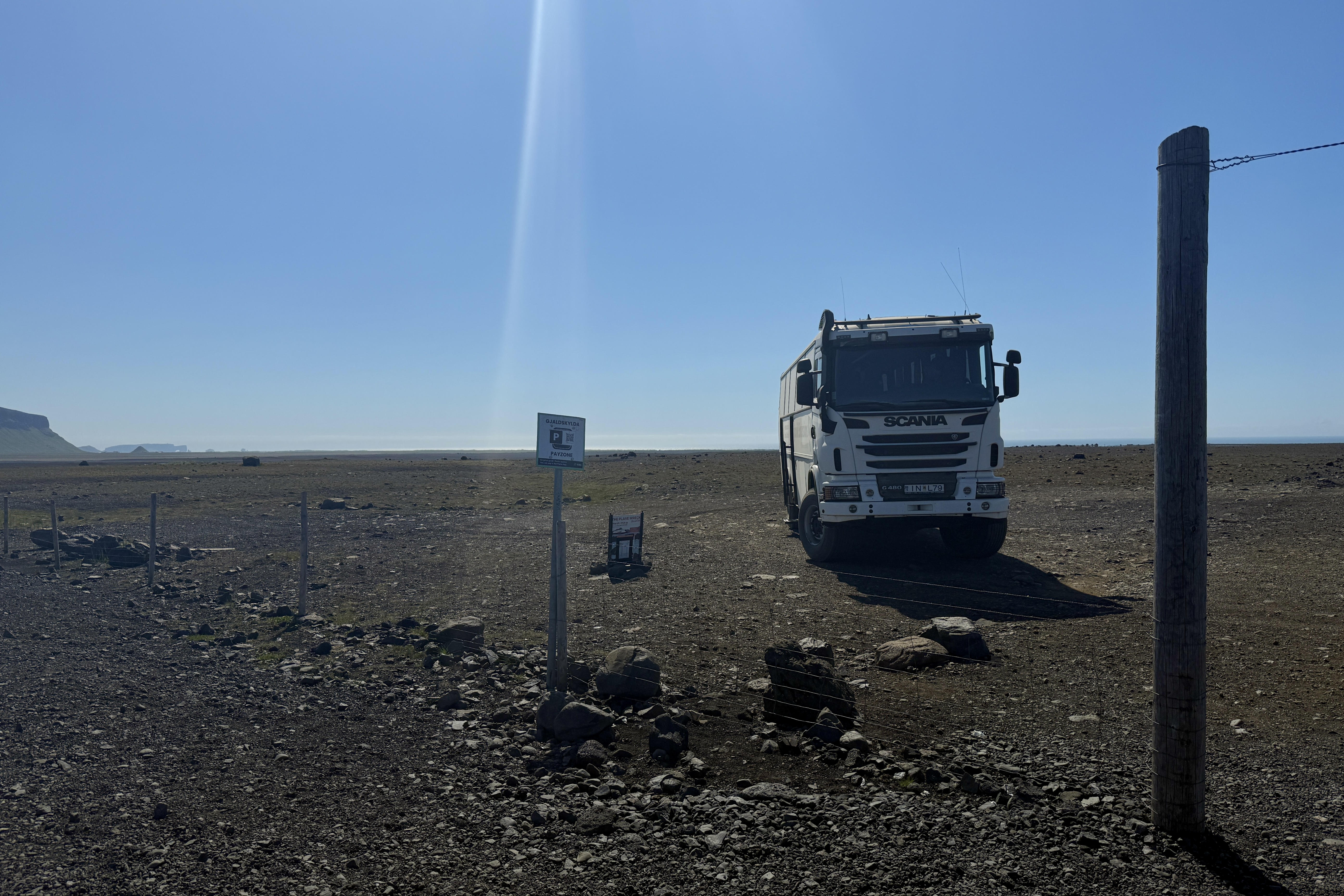 검은 화산재 지형으로 이루어진 황무지에 셔틀버스가 세워져 있고, 왼쪽에는 작은 표지판이 있습니다. 주변에는 돌과 모래만 있는 황량한 풍경이 펼쳐져 있으며, 배경에는 푸른 하늘과 멀리 바다가 보입니다.
