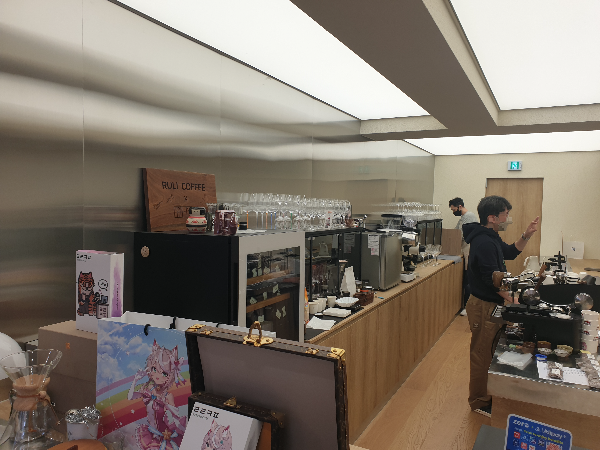 바리스타 두 명이 일하고 있는 모습. 우드 인테리어와 흰색 벽면과 스틸로 된 작업 벽면 공간이 보인다.