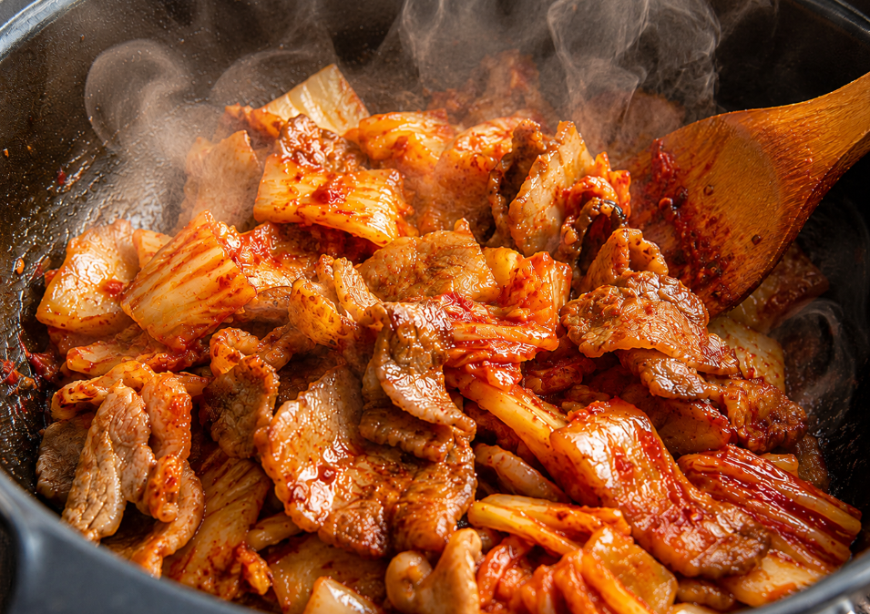 달궈진 냄비 안에서 붉은 김치와 돼지고기가 들기름에 달달 볶아지며 맛있는 향을 내는 조리 장면.