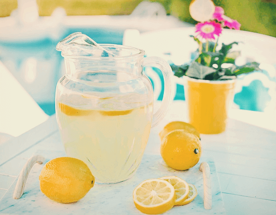 투명한 유리 주전자에 레몬이 담긴 레몬즙이 가득하고 주변엔 노란 레몬들이 있다.